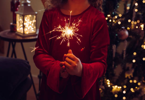 criança menina bonita segurando vela cintilante nas mãos na sala de estar de casa, árvore de natal decorada ao fundo, quarto escuro, iluminada por vela cintilante. saudações felizes de ano novo. - sparkler sparks new years eve human hand - fotografias e filmes do acervo