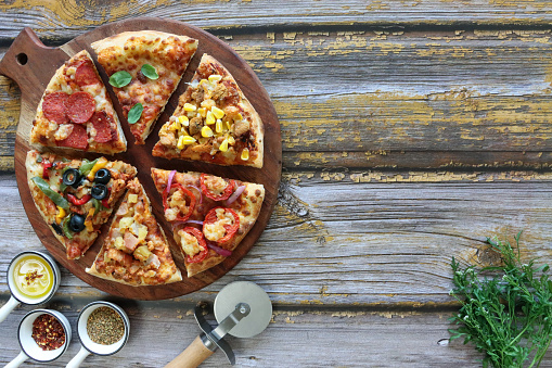 Stock photo showing elevated view of circular, wooden chopping board containing triangular slices of pizzas with different toppings including Hawaiian pizza (pineapple and ham), chicken and sweetcorn, pepperoni, vegetable (yellow and green bell pepper with black olives), feta, tomato and red onion, and Pizza Margherita (melted golden buffalo mozzarella cheese, rich tomato marinara sauce and fresh basil leaves).