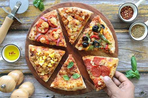 Stock photo showing close-up, elevated view of circular, wooden chopping board containing triangular slices of pizzas with different toppings including Hawaiian pizza (pineapple and ham), chicken and sweetcorn, pepperoni, vegetable (yellow and green bell pepper with black olives), feta, tomato and red onion, and Pizza Margherita (melted golden buffalo mozzarella cheese, rich tomato marinara sauce and fresh basil leaves).