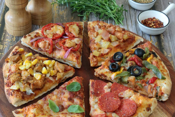 Stock photo showing elevated view of circular, wooden chopping board containing triangular slices of pizzas with different toppings including Hawaiian pizza (pineapple and ham), chicken and sweetcorn, pepperoni, vegetable (yellow and green bell pepper with black olives), feta, tomato and red onion, and Pizza Margherita (melted golden buffalo mozzarella cheese, rich tomato marinara sauce and fresh basil leaves). imagen de primer plano rebanadas de pizza, pizza hawaiana, feta, rodajas de tomate y cebolla roja, pollo y maíz dulce, pepperoni, pizza margherita, verduras y aceitunas negras, tabla de cortar de madera, rueda de pizza, molinos de sal y pimienta, fondo de