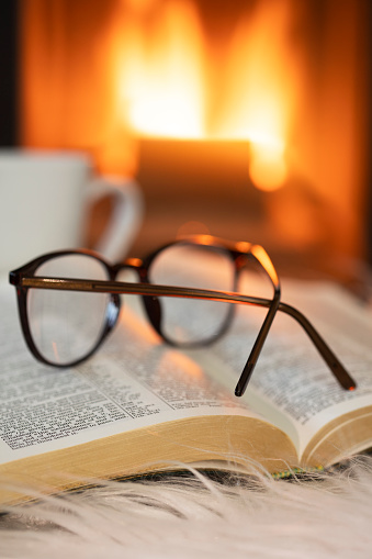Open book with eyeglasses and  cup of tea in front of the fireplace