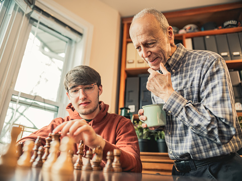 Grandfather and Grandson playing chess at home. Both dressed in casual clothes and grandson wearing eyeglasses. Sitting at the chess table by the window. Interior of private home in winter time.