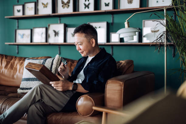 istock Senior Asian man relaxing on sofa at cozy home, examining insect collection specimens with magnifying glass. Happy retirement life, hobbies and interests for the elderly concept 1360541242