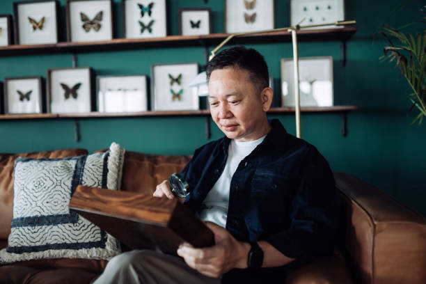 istock Senior Asian man relaxing on sofa at cozy home, examining insect collection specimens with magnifying glass. Happy retirement life, hobbies and interests for the elderly concept 1360541277