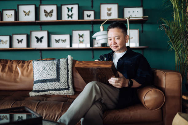 istock Senior Asian man relaxing on sofa at cozy home, examining insect collection specimens with magnifying glass. Happy retirement life, hobbies and interests for the elderly concept 1360541377