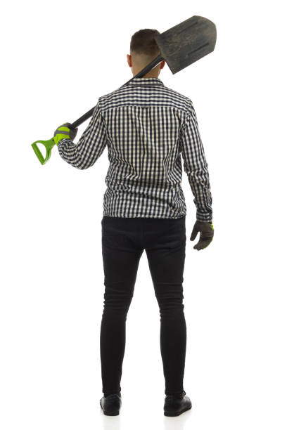 Man in lumberjack shirt, gloves and black jeans is standing with spade on his shoulder. Rear view. Full length studio shot isolated on white.