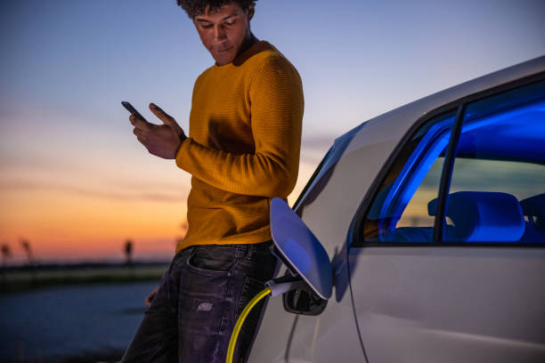 man using mobile phone while charging electric car - electric car imagens e fotografias de stock