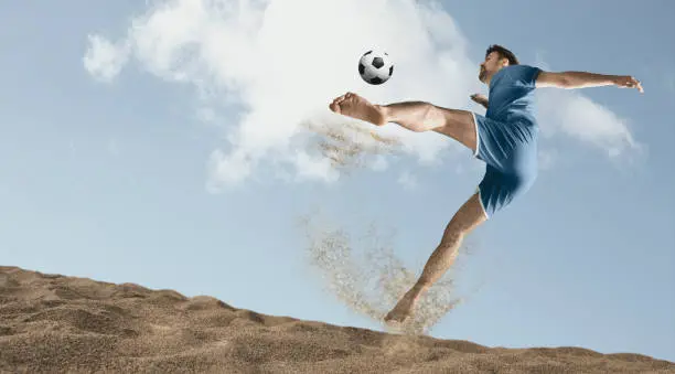 The man footballers are desperately playing beach soccer on sand on a sunny day. Man doing kick on beach with soccer ball