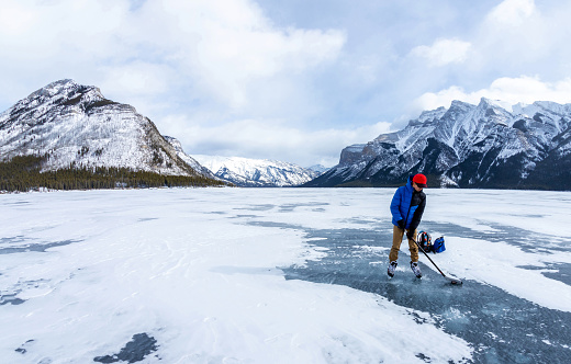 Boy practicing ice hockey on frozen Lake Minnewanka in the Canadian Rockies of Banff National Park during the cold Winter with unidentifiable visitors walking on the lake in the background.