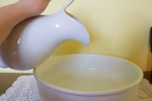 A woman's hand pours water from a white ceramic water jug ​​into a ceramic vessel.
