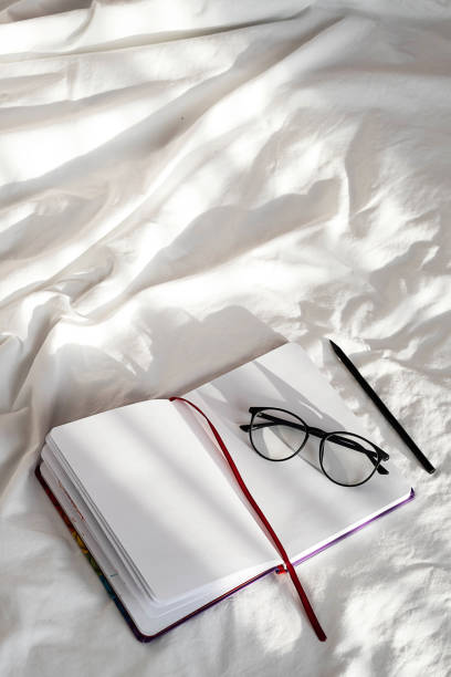 istock Diary, pencil and eyeglasses rest on white bedding in sunlight with shadows.  Personal journal for notes. Stationary still life scene, morning at home. 1360619769
