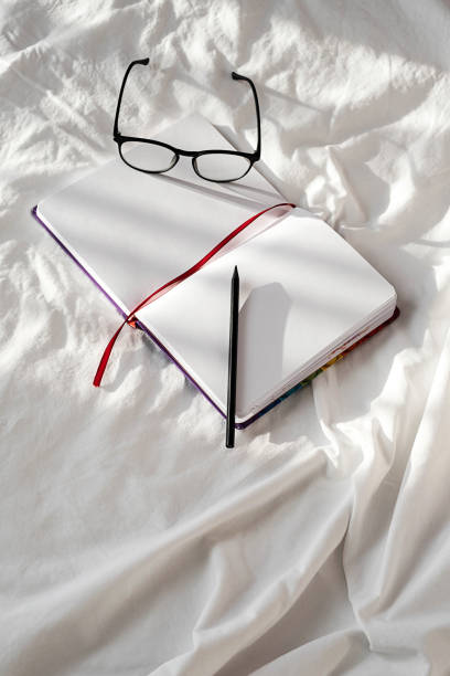 istock Blank notebook, pencil and eyeglasses rest on white bedding in sunlight with shadows. Stationary still life scene of home office, copy space. Overhead shot, minimal style. 1360619770