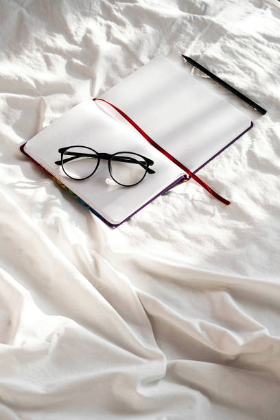 istock Diary, pencil and eyeglasses rest on white bedding in sunlight with shadows.  Personal journal for notes. Stationary still life scene, morning at home. 1360619771