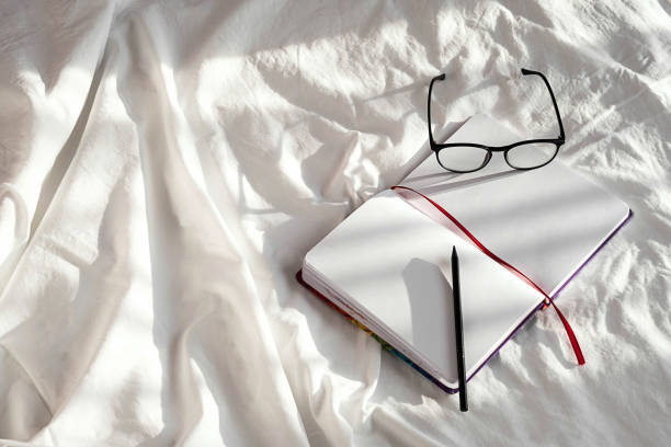 Blank notebook, pencil and eyeglasses rest on white bedding in sunlight with shadows. Stationary still life scene of home office, copy space. Overhead shot, minimal style.