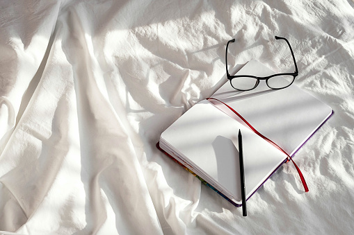 Blank notebook, pencil and eyeglasses rest on white bedding in sunlight with shadows. Stationary still life scene of home office, copy space. Overhead shot, minimal style.