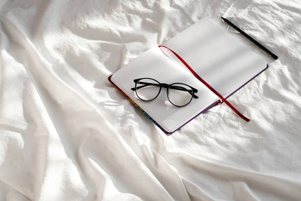 istock Diary, pencil and eyeglasses rest on white bedding in sunlight with shadows.  Personal journal for notes. Stationary still life scene, morning at home. 1360619779