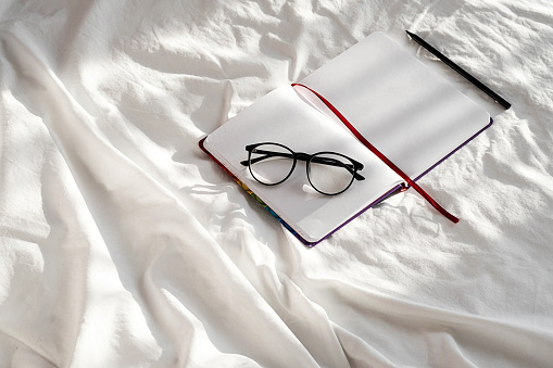 Diary, pencil and eyeglasses rest on white bedding in sunlight with shadows.  Personal journal for notes. Stationary still life scene, morning at home.