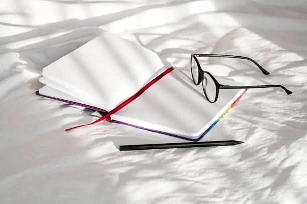 istock Blank notebook, pencil and eyeglasses rest on white bedding in sunlight with shadows. Stationary still life scene of home office, copy space. Overhead shot, minimal style. 1360619780