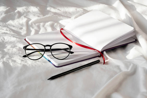 istock Diary, pencil and eyeglasses rest on white bedding in sunlight with shadows.  Personal journal for notes. Stationary still life scene, morning at home. 1360619787