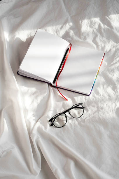 istock Blank notebook, pencil and eyeglasses rest on white bedding in sunlight with shadows. Stationary still life scene of home office, copy space. Overhead shot, minimal style. 1360619792