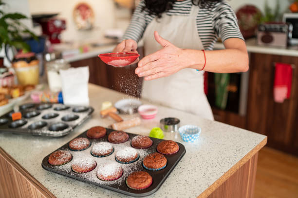 Woman's hands, using strainer to sprinkle powdered sugar at freshly backed homemade muffins in a tray.