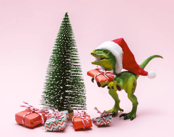 istock Funny plastic toy dinosaur wearing Santa Claus hat with  present boxes on a pink background. 1360649004