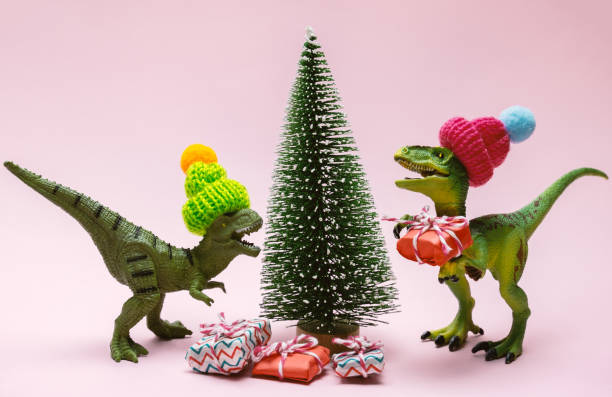 istock Funny plastic toy dinosaurs wearing knitted hat with  present boxes and evergreen tree on a pink background. 1360649010