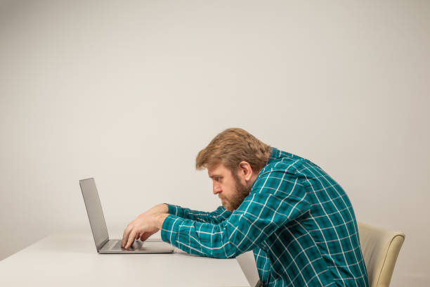 hombre emocional de barba en posición encorvada sentado en la sala de oficina, trabajando con una computadora portátil - modo de vida no saludable fotografías e imágenes de stock