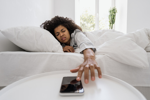 Tired and unhappy afro american woman reach hand to dismiss or snooze alarm clock on her blank screen smartphone. Female dont want wake up at early morning. Millennial girl in sleepwear lying on bed