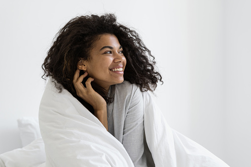 Happy dark skinned woman wrapped in a soft blanket, looking aside, spending early morning in bedroom. Cheerful millennial girl wearing in sleepwear, waking up, enjoying lazy weekend day at home