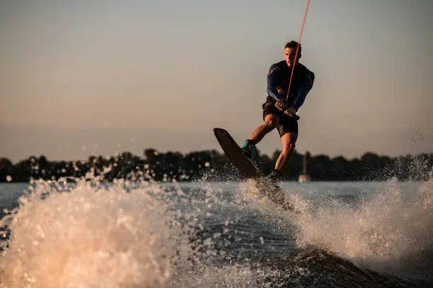 athletic male wakeboarder holding rope and jumps with wakeboard over splashing river. Summertime watersports activity
