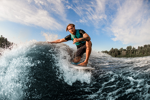 athletic guy wakesurfer skilfully riding down the blue splashing wave on a warm day. Active water sports