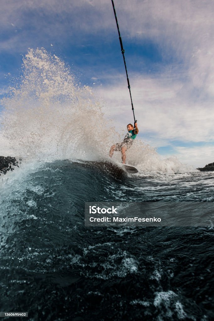 view of great splashing wave and man holds rope and glides on the waves on the wakesurf board view of great splashing wave and man holds rope and glides on the waves on the wakesurf board on the background of sky Wakeboarding Stock Photo