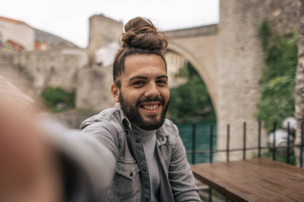 istock Young bearded man sitting on bench and making selfie with arch bridge in background 1360696616