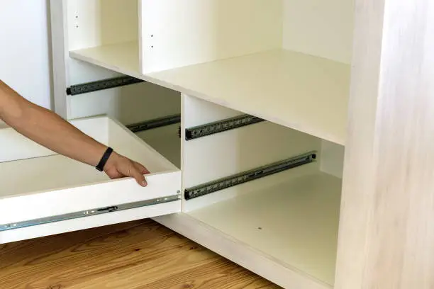 Photo of Close up of carpenter hands installing wooden drawer on sliding skids in contemporary cupboard cabinet.