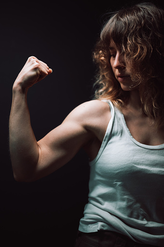 Close-up of a young woman showing her biceps and strength on a dark background.