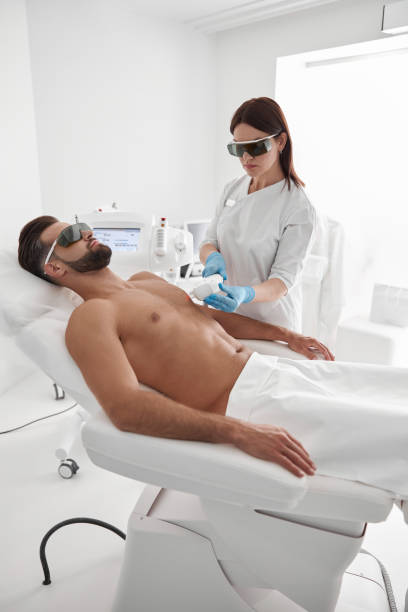 istock Young master with goggles conducts laser epilation to bearded man client lying on couch in clinic 1360716184