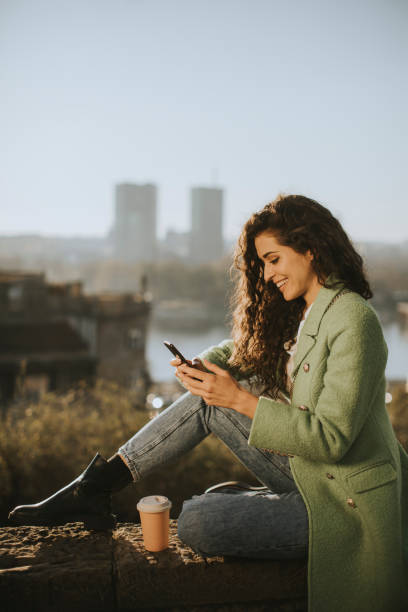 istock Pretty young woman using smartphone while sitting by the river and drinking takeaway coffee 1360718205