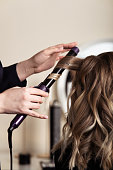 istock Female hands of a hair master. Close-up of blond hair on a curling iron. Curling curls. 1360736579