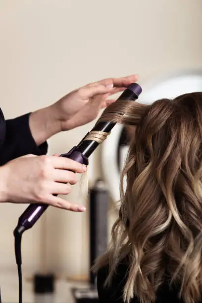 Photo of Female hands of a hair master. Close-up of blond hair on a curling iron. Curling curls.