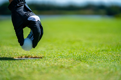 Close up hand of Asian senior man golfer picking golf ball out of the hole on green at golf course. Healthy male enjoy outdoor lifestyle activity sport golfing at country club on summer vacation