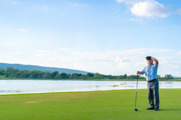 Asian senior man holding golf club hitting golf ball on fairway at country club in sunny day. Healthy elderly male golfer enjoy outdoor activity sport golfing at golf course on summer vacation