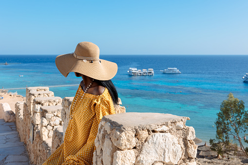Happy African American woman in yellow dress and sun hat enjoys view of coast of Red Sea on natural background. Panoramic views of blue sea with yachts and coastline, Sharm El Sheikh, Egypt.