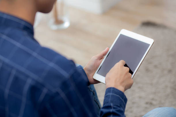 istock Closeup young asian man sitting and using mockup of tablet computer with display blank screen on sofa at home, male holding digital tablet watch entertainment at living room, communication concept. 1360757994