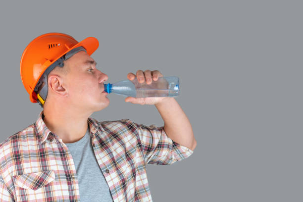 istock A tired worker drinks water from a plastic bottle standing on a gray background, copyspace 1360768105