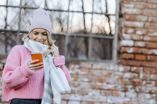 Cheerful young woman texting on mobile phone in front of brick wall in the city