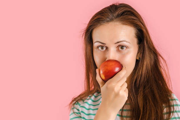 istock Cute brunette vegetarian sniffs a red apple standing on a pink background, copyspace 1360780719