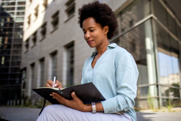 istock Young African American woman writing in a small notepad journal 1360786015