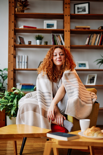 A happy redhead girl covered with a blanket is sitting in her armchair in the living room and enjoying morning moments.