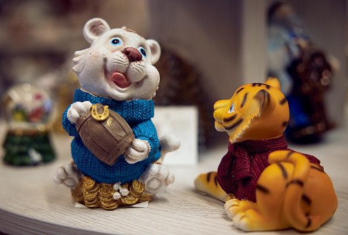 New Year's toys. Figurine white lion cub piggy bank with a barrel of money in his hands in a blue sweater and a figurine of a red lion cub in a red sweater on a shelf in a supermarket.